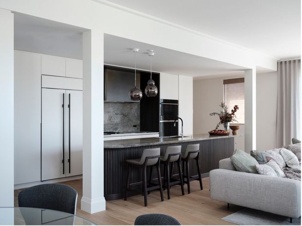 Open-plan contemporary kitchen and living area with a large island and integrated breakfast bar. The kitchen features flat-panel white cabinetry, dark vertically ribbed island joinery, a stone benchtop and splashback, black appliances and plumbing fixtures, and metallic globe pendant lights. A light upholstered sectional sofa and dining area are visible in the adjoining space, with soft neutral finishes and abundant natural light.