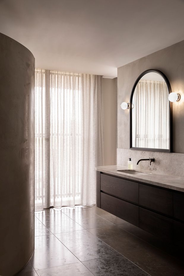 A refined contemporary bathroom with a dark floating timber vanity, pale stone benchtop and integrated basin, wall-mounted black tapware, and a large arched black-framed mirror. Soft globe wall lights flank the mirror. Full-height sheer curtains filter daylight at the far end, while large dark grey stone-look floor tiles and warm taupe plastered walls create a calm, moody palette.