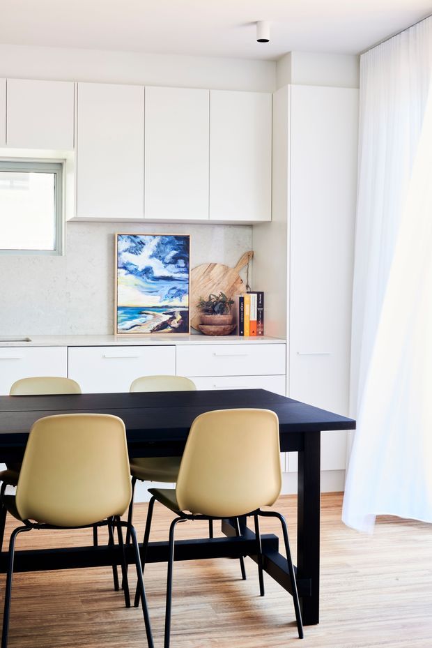 Bright, minimalist dining area with a black rectangular dining table and four pale yellow upholstered chairs. White floor-to-ceiling cabinetry lines the rear wall, with a light stone-look backsplash, open shelving displaying artwork and decor, a small window, sheer white curtains, and warm timber flooring.