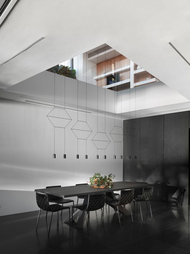 Contemporary dining room with a long dark dining table and woven black dining chairs beneath a dramatic linear arrangement of geometric pendant lights. The room features dark floor tiles, a black built-in wall, crisp white walls and ceiling, and a large ceiling opening revealing an upper-level space with timber joinery and indoor planting.