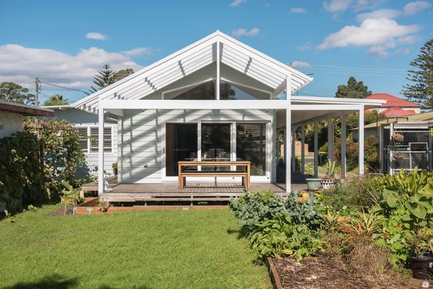 Front exterior view of a compact single-storey house with a prominent white gabled roof, horizontal weatherboard cladding, exposed covered porch structure, and large glazed openings. The home opens onto a lawn and landscaped garden under a bright blue sky, with neighbouring residential structures visible in the background.