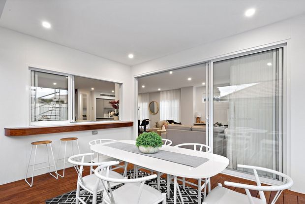 Bright contemporary dining area with a white oval dining table and matching white chairs, set on a black-and-white patterned rug. The room features warm timber flooring, white walls and ceiling with recessed downlights, a timber pass-through ledge with stools, and large sliding glass doors opening to a courtyard or outdoor area. A living room is visible beyond the glazing.