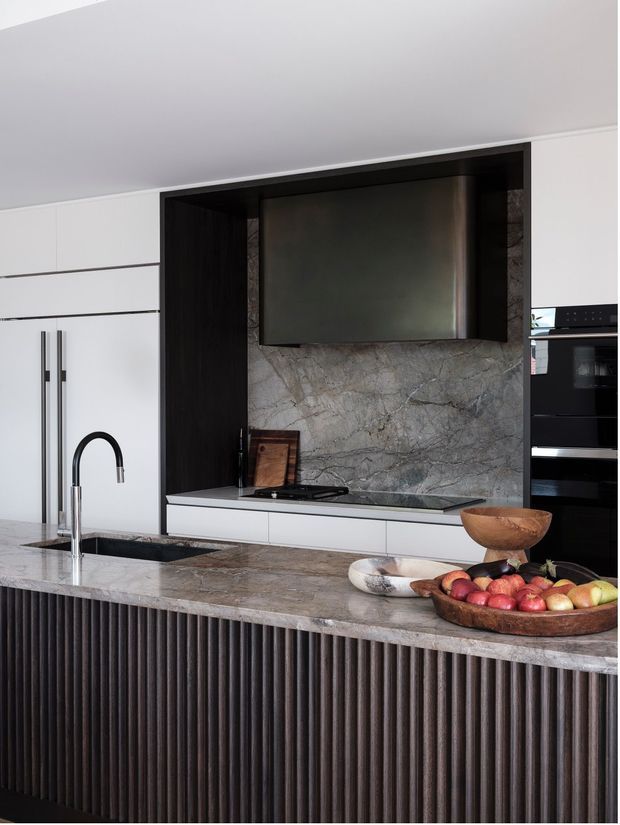 A sophisticated contemporary kitchen featuring white full-height cabinetry, dark timber-toned fluted island joinery, a large veined stone island benchtop, matching stone splashback, integrated black appliances, and a sculptural black gooseneck tap. The island is styled with wooden bowls and fruit.