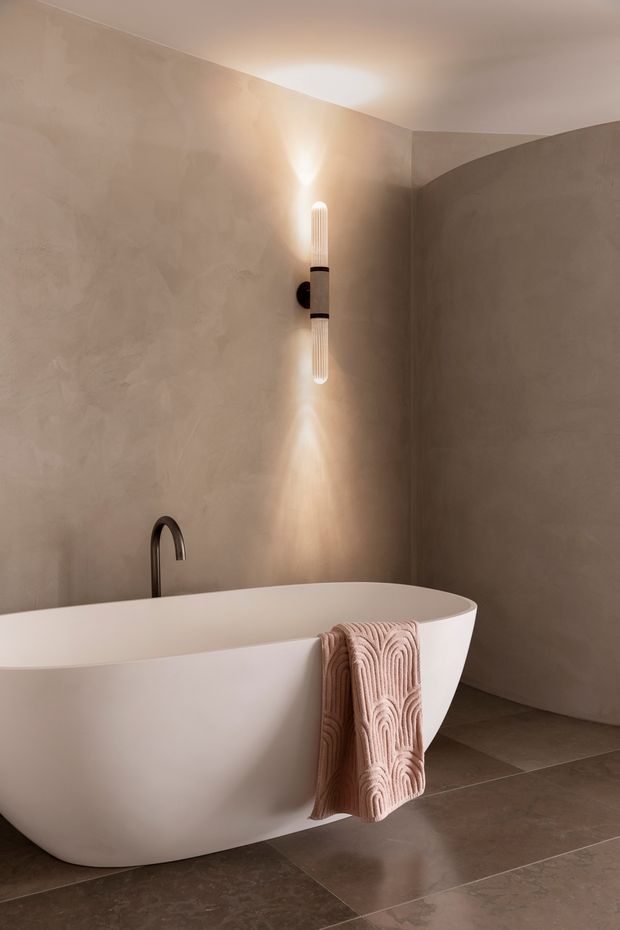 A serene, minimalist bathroom featuring a freestanding white bathtub with a dark curved faucet, set against softly textured taupe plaster walls. A vertical ribbed wall sconce casts warm upward and downward light, while large taupe stone-look floor tiles and a folded blush-toned towel create a warm, refined atmosphere.