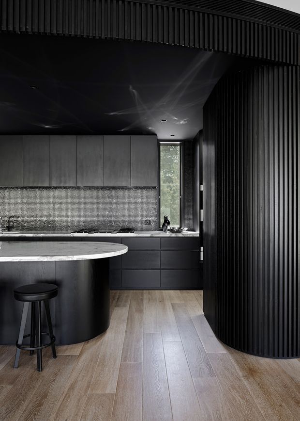 A sophisticated dark-toned kitchen with a curved island featuring a pale stone benchtop, black curved cabinetry, integrated dark drawers, and a shimmering small-format mosaic splashback. Vertical black slatted wall and ceiling detailing creates a dramatic architectural enclosure, while pale timber-look plank flooring and a narrow window introduce contrast and natural light. A single black bar stool sits at the island.