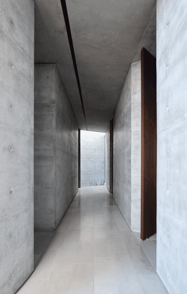 A narrow minimalist interior corridor with exposed smooth concrete walls and ceiling, a pale tiled floor, and a dark timber doorway on the right. The hallway leads toward a bright glazed opening at the far end, creating a strong linear perspective and understated architectural atmosphere.