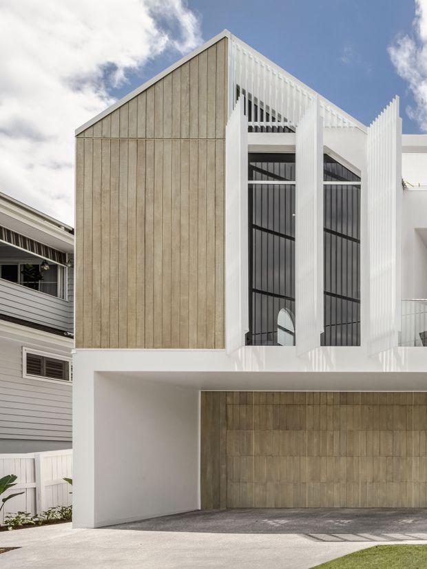 Street-facing view of a contemporary multi-storey residence with a steep gable-like facade, white structural framing, tall dark-framed glazing, and vertical timber-look cladding. A wide integrated garage occupies the lower level, with neighbouring residential buildings visible at the sides.