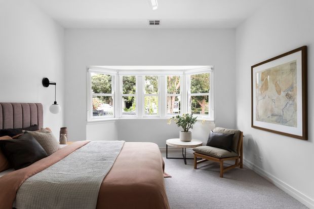 A bright, calm bedroom with a large multi-panel bay window, soft white walls, pale carpet, and a muted blush upholstered bed. A sculptural black wall lamp, small round side table, upholstered timber-framed lounge chair, potted plant, and framed artwork complete the refined contemporary interior.