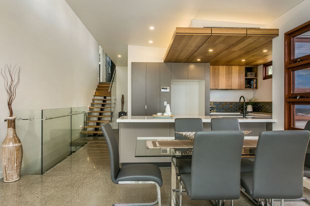 Open-plan contemporary kitchen and dining area with a polished concrete floor, sleek grey cabinetry, a large island, and a timber-lined ceiling canopy extending over the kitchen. A glass balustrade borders the stairway, while a dark-framed window and patterned tile splashback add contrast. Grey upholstered dining chairs occupy the foreground.