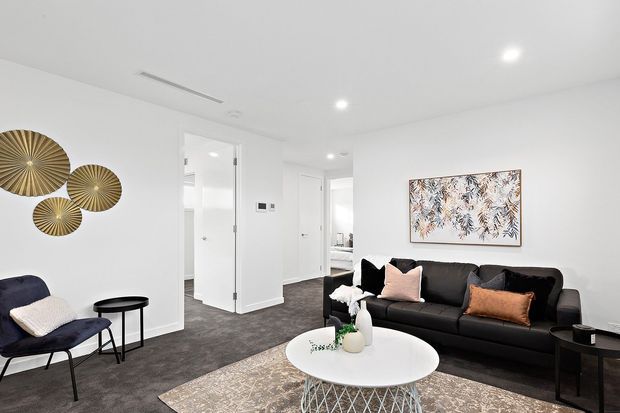 Bright contemporary living room with white walls and ceiling, dark carpet flooring, a black sofa with blush and rust cushions, a navy accent chair, round nesting side tables, and a white round coffee table. Decorative gold-toned wall discs and botanical artwork add contrast, while open doorways lead to adjacent spaces.