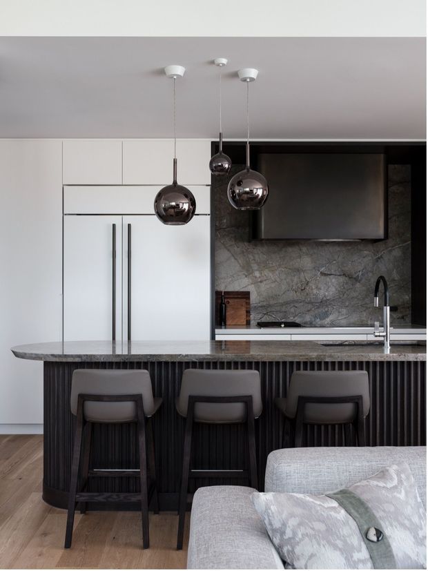 A refined contemporary kitchen viewed from the living area, featuring a large dark stone island with integrated seating, vertically fluted dark cabinetry, white full-height cabinetry, a dramatic veined stone splashback, black rangehood, and reflective spherical pendant lights. A pale upholstered sofa is visible in the foreground.