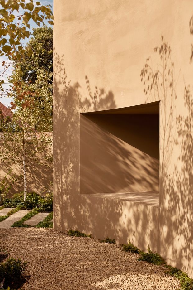 A minimalist architectural exterior featuring a tall warm beige rendered wall with a deeply recessed rectangular opening. Strong sunlight creates dramatic diagonal illumination and crisp botanical shadows across the facade. A gravel ground plane, stepping-stone path, low planting, and mature trees frame the building, with a muted residential roofline visible in the background.
