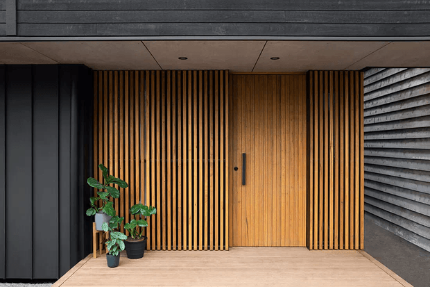 A contemporary residential entryway with a warm timber slatted feature wall and flush timber door, framed by dark vertical cladding and a textured grey side wall. Recessed ceiling lights, timber flooring, and potted greenery create a refined, welcoming entrance.