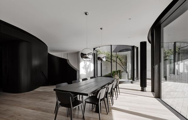 A refined contemporary dining area with a long dark dining table and black chairs, positioned within a bright open-plan interior. The room features dramatic curved black ribbed wall and joinery forms, polished spherical pendant lights, pale timber flooring, extensive floor-to-ceiling glazing, and a planted courtyard visible beyond the glass. Strong contrast between the dark architectural elements and abundant natural light creates a sophisticated, sculptural atmosphere.