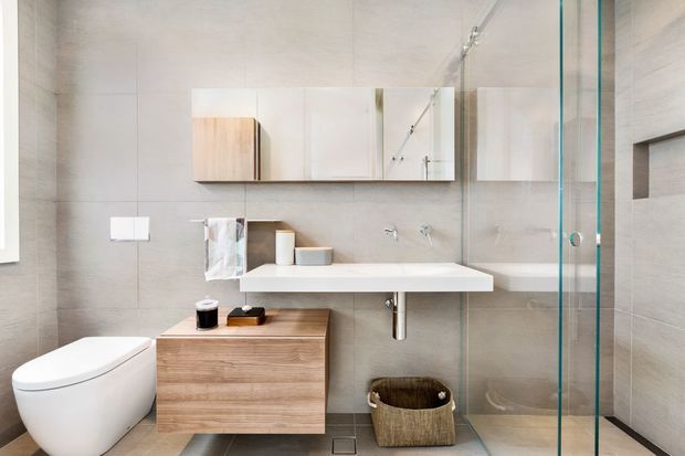 Contemporary minimalist bathroom with a wall-mounted timber vanity, white integrated basin, mirrored cabinet, wall-hung toilet, large-format grey wall and floor tiles, and a frameless glass shower enclosure. The room is bright, clean, and highly streamlined with warm wood accents.