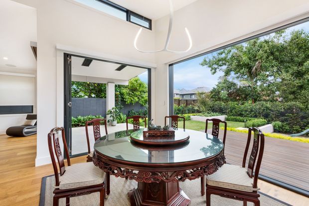Bright dining room with a large ornate round timber dining table and carved wooden chairs, opening onto a timber deck and landscaped garden through expansive glazing. The room has pale walls, timber flooring, a contemporary sculptural pendant, and a view toward neighbouring homes and greenery.
