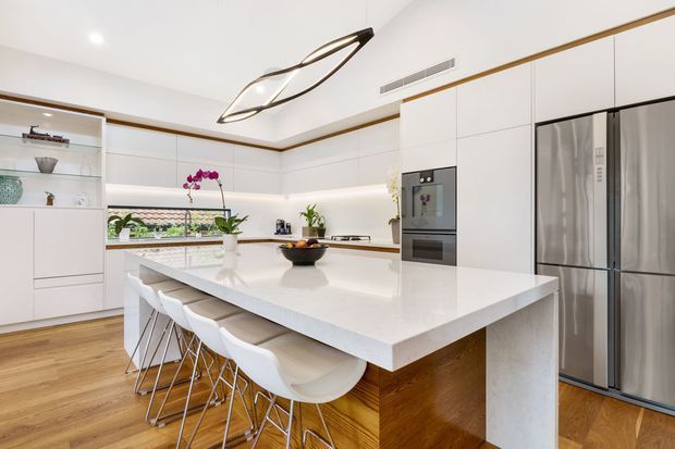 Bright, spacious contemporary kitchen with extensive white flat-panel cabinetry, a large white island with waterfall edge and integrated seating for five, timber veneer island sides, warm timber flooring, stainless-steel appliances, open shelving, and a sculptural modern pendant light. The room has a clean, refined, high-end feel with subtle warm timber accents.