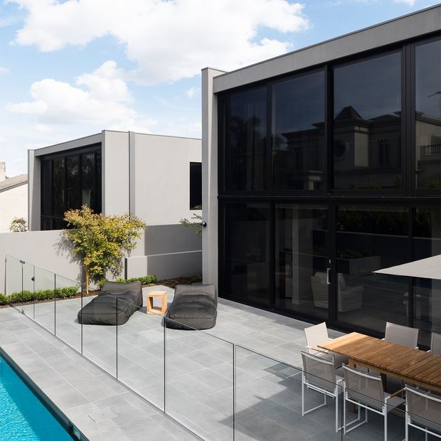 A contemporary multi-level residential courtyard terrace viewed from above, with large black-framed floor-to-ceiling glazing, smooth light rendered walls, and a broad grey tiled deck. The terrace includes a dining table, informal lounge seating, frameless glass pool fencing, and a swimming pool visible at the edge. Bright daylight and strong geometric lines create a clean, modern outdoor entertaining space.