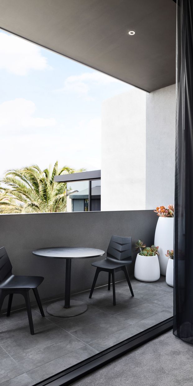 A contemporary covered balcony or terrace viewed from inside through full-height dark-framed glazing. The outdoor space has charcoal rendered walls, a smooth grey tiled floor, a slim round black café table, black chairs, and white planters with succulents. A palm tree and neighbouring modern architecture are visible beyond the low parapet. The ceiling is a dark smooth soffit with a recessed downlight, and full-height dark curtains frame the opening.