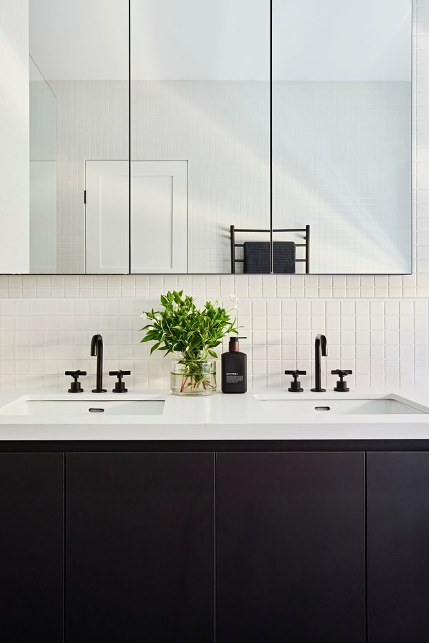A bright, symmetrical contemporary bathroom vanity with two white integrated basins, matte black tapware, dark flat-front cabinetry, and a large frameless mirror. White square wall tiles form the backsplash, with a small plant and soap dispenser on the countertop. The mirror reflects a white door and heated towel rail.