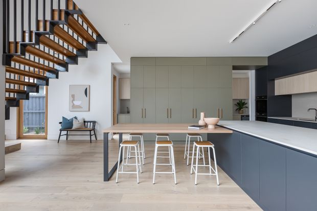 Contemporary open-plan kitchen and dining area with a large island and breakfast bar, pale timber flooring, muted green-grey cabinetry, dark navy lower cabinetry and wall panels, and a floating timber-and-black-metal staircase with open risers. The space is bright and refined, with a small seating nook and garden glimpsed through the glazing.