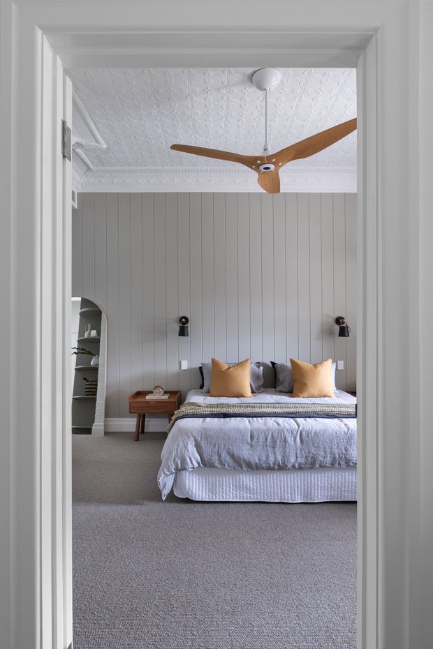 A calm, refined bedroom viewed through a white-trimmed doorway. The room features a vertically panelled grey wall, an ornate pressed-pattern white ceiling with decorative cornice, a timber three-blade ceiling fan, wall-mounted bedside lights, a neatly made bed with white and grey bedding and ochre accent cushions, grey carpet, and built-in arched shelving at the side.