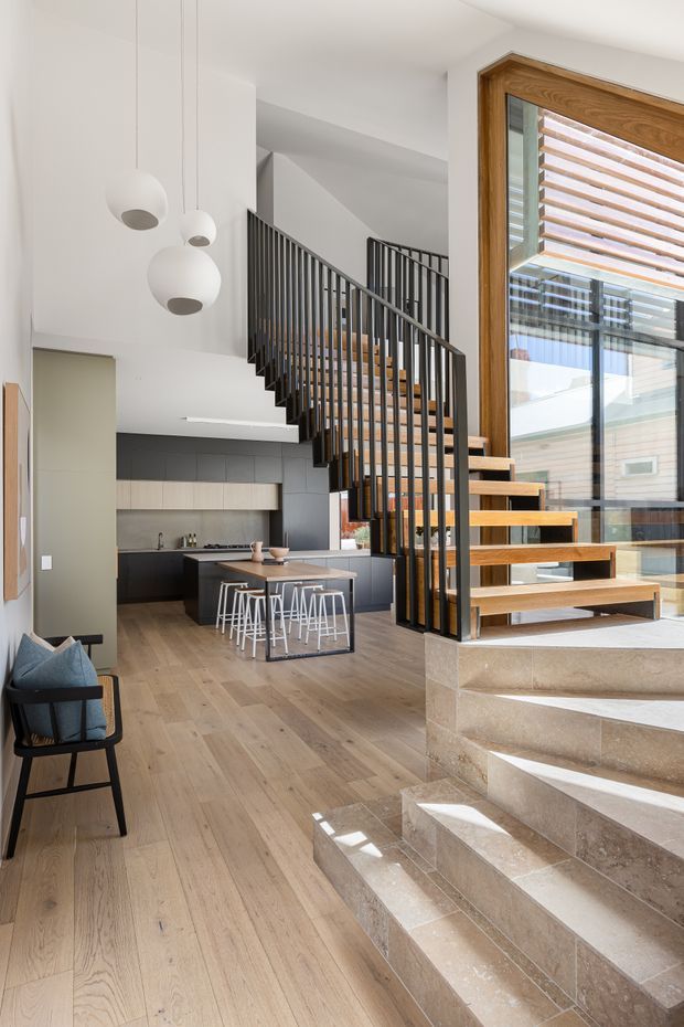 A bright, contemporary open-plan interior featuring a timber staircase with black metal balustrades, pale timber flooring, a dark kitchen with a central island and white bar stools, and large timber-framed glazing with external horizontal screening. Stone-clad steps lead up through the space, while spherical pendant lights hang over the kitchen and dining area.