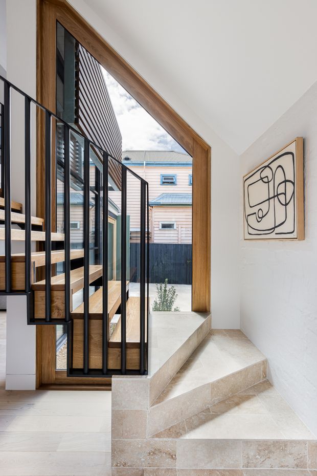 Interior view of a compact stairway and landing with pale stone-tiled steps, light timber flooring, a sculptural timber-framed opening, and black metal balustrades. White walls and ceiling create a bright, clean setting, with a framed abstract line artwork on the wall and an exterior courtyard visible through the opening.