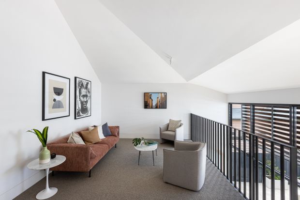 Bright contemporary living area on an upper-level landing or mezzanine, furnished with a rust-coloured sofa, armchairs, round coffee table and small side table. White walls and an angled ceiling create an airy space, with framed artwork and a large louvred window beside a black metal balustrade overlooking the level below.