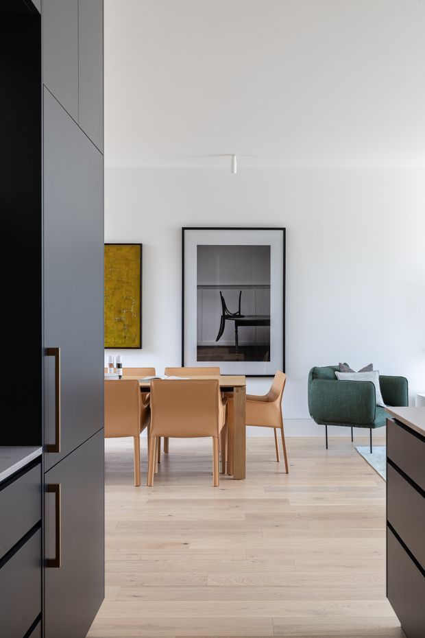 A bright, minimalist open-plan interior viewed from a dark built-in kitchen or cabinetry zone. The scene centers on a timber dining table with tan leather dining chairs, opening into a living area with a muted green upholstered armchair. Pale timber flooring, white walls, dark cabinetry, framed artwork, and restrained contemporary furnishings create a refined modern look.
