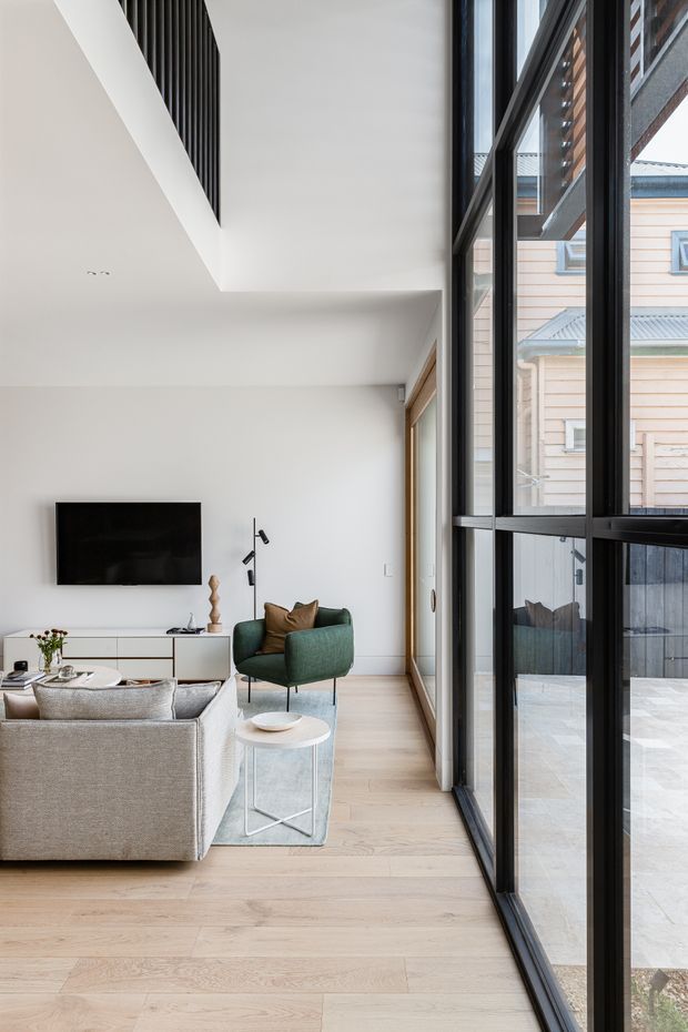 Bright contemporary living room with pale timber flooring, white walls, a large sectional sofa, green armchair, wall-mounted television and a slim round side table. Full-height black-framed glazing runs along the right side, looking onto a paved courtyard and neighbouring building. The room has a high ceiling with a narrow dark vertical feature near the upper wall.
