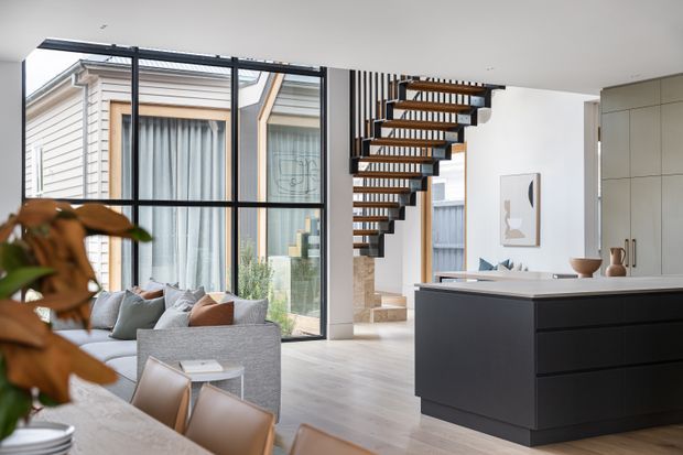 Bright contemporary open-plan interior featuring a kitchen island with dark built-in cabinetry, a living area with a grey sofa, large black-framed glazing, and a visually prominent open-riser timber staircase. Light timber flooring, white walls, and a compact dining area create an airy, refined space.