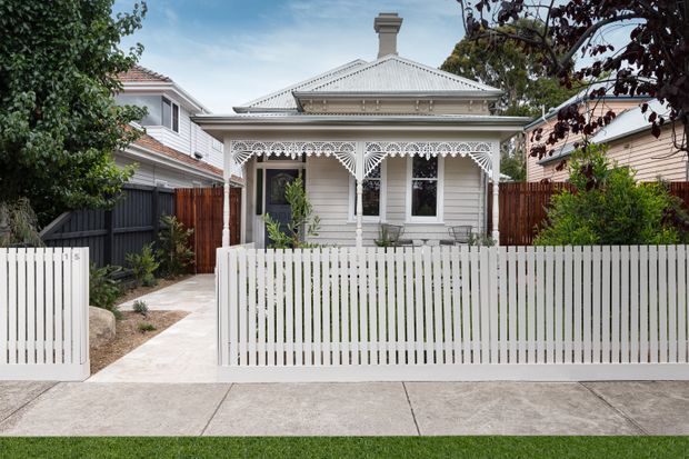 Front exterior view of a traditional single-storey cottage with a pale weatherboard facade, hipped corrugated metal roof, decorative lacework verandah, white picket fence, timber gates, and a paved front path. The home is set in a compact leafy suburban garden.