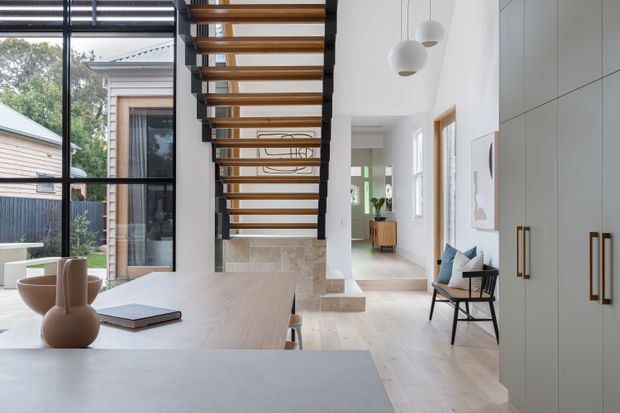 Bright contemporary open-plan interior featuring a light timber dining table in the foreground, a sculptural open-riser timber staircase with dark stringers, floor-to-ceiling black-framed glazing, pale stone stair and wall finishes, and tall built-in cabinetry with brass-toned handles. A reading chair and hallway are visible beyond, with pendant lights adding a refined accent.
