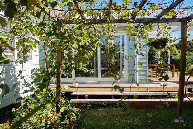 A leafy outdoor view of a timber pergola or trellis covered with climbing vines, framing a raised timber deck and a light-coloured weatherboard house. Large sliding glazed doors open onto the deck, where a wooden outdoor dining table and chairs are visible. The garden setting is sunny and green, with a hanging planter and dense planting around the structure.
