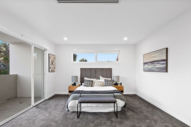 A bright contemporary bedroom with a centered bed, charcoal upholstered headboard, layered neutral bedding, timber bedside tables, and a black metal bench. The room has white walls and ceiling, grey carpet, recessed lighting, a high horizontal window, and large sliding glass doors opening to an exterior balcony or terrace.