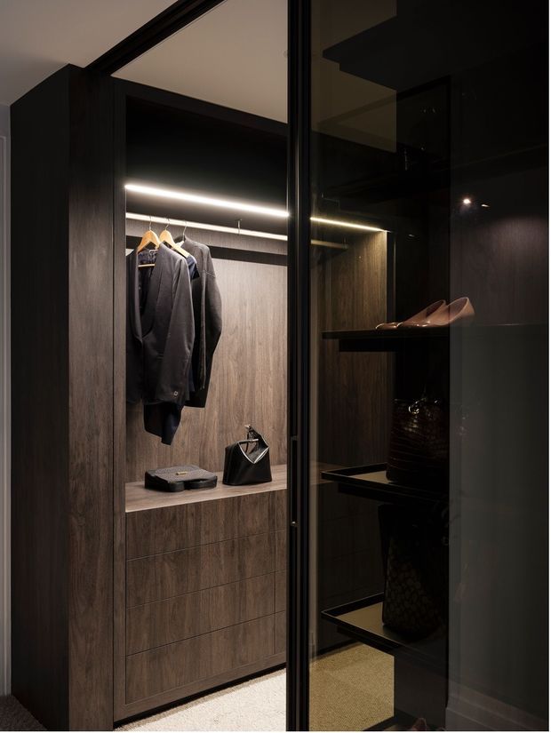 A dark, refined walk-in wardrobe with floor-to-ceiling built-in timber-look cabinetry, hanging rails, drawers, open shelves, and integrated linear LED lighting. Tailored clothing, shoes, handbags, and accessories are arranged within the joinery, with black-framed glass doors and reflective surfaces creating depth. The atmosphere is moody and luxurious.