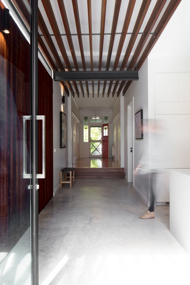 A bright contemporary residential entry hall viewed through a large glazed front door. The space has a polished concrete floor, white walls and built-in cabinetry, a dark timber feature wall and door, and a striking ceiling of regularly spaced exposed timber battens with dark structural steel framing. A short set of timber steps leads toward a glazed rear door, with artwork and a small bench along the corridor.