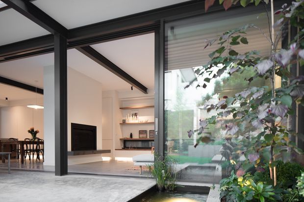 A contemporary indoor-outdoor living space viewed from a landscaped courtyard. Black steel-framed glazing and structural posts open onto a bright living room with a fireplace, built-in shelving, pale furnishings, and an adjacent dining area. A shallow reflecting pond and dense planting occupy the foreground, creating a strong connection between the interior and garden.