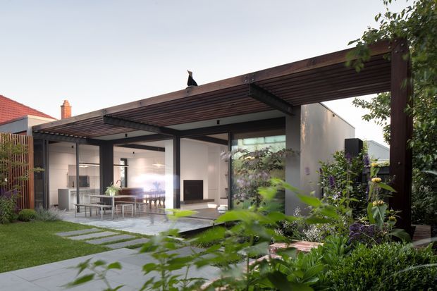A landscaped backyard courtyard viewed from the garden, featuring a contemporary single-storey pavilion or extension with extensive floor-to-ceiling glazing, dark timber pergola-like roof framing, and a pale rendered wall. Inside, a dining table and chairs are visible beside a minimalist living area with a fireplace. Stepping stones cross the lawn toward a paved terrace, while dense planting surrounds the structure.