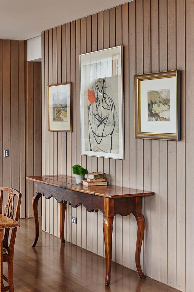 A warmly styled interior vignette featuring a narrow antique console table with ornate scalloped apron and curved legs against a full-height vertical timber-panelled wall. Three framed artworks hang above the table, which is accessorised with stacked books and a small potted plant. A partial carved wooden chair is visible at left, and the polished timber floor complements the rich brown wood palette.