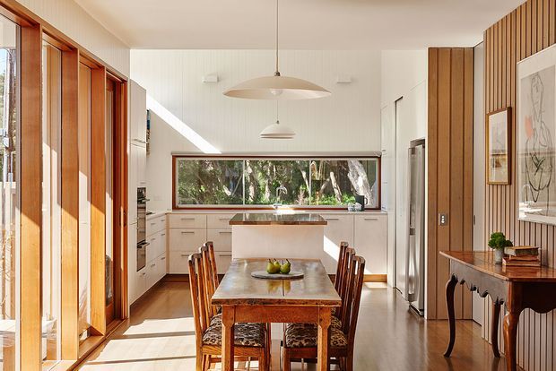 A bright, warm kitchen and dining area viewed from the living side. The open-plan space features extensive timber-framed glazing and doors on the left, a white galley-style kitchen with a central island, a long horizontal window overlooking trees, timber flooring, and a timber dining table with traditional carved chairs. One wall is clad in vertical timber battens, with pendant lights and a dark traditional console adding character.