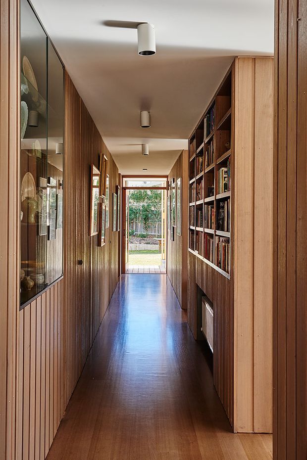 A narrow, long residential hallway lined with warm vertical timber wall panelling and integrated joinery. A glass-fronted display cabinet occupies the left wall, while open built-in bookshelves run along the right. The polished dark timber floor reflects a series of simple cylindrical ceiling lights, and a glazed doorway at the far end opens toward a garden. The space has a refined, warm, library-like character with strong linear perspective.