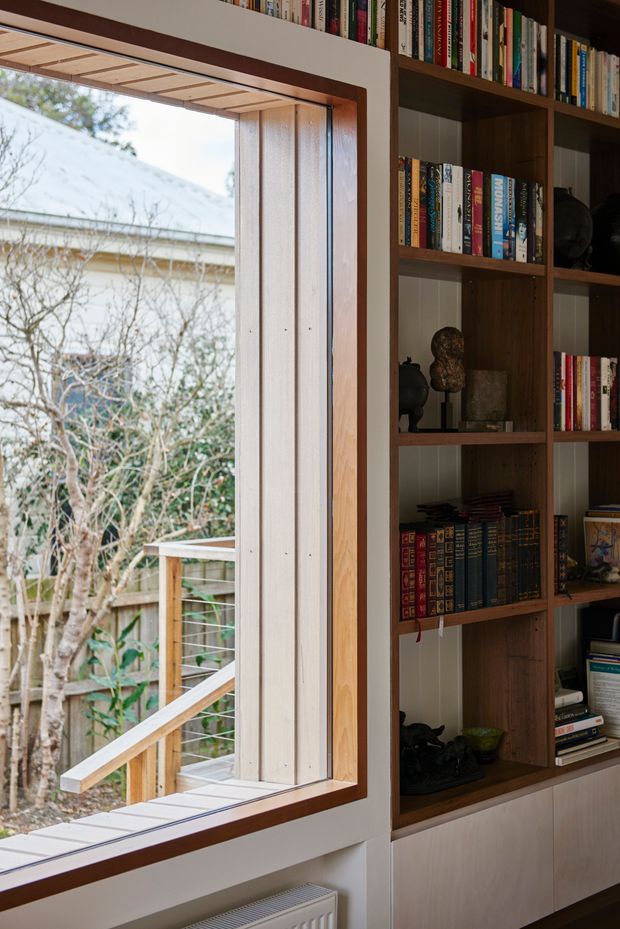 A close-up interior view of a timber-framed window beside a full-height built-in bookcase. The window has pale vertical timber reveals and a deep dark timber trim, looking onto a small deck and garden. The shelving is dark-stained timber with books and decorative objects, set above light-toned lower cabinetry. The space feels warm, refined, and intimate, with a strong connection to the outdoors.