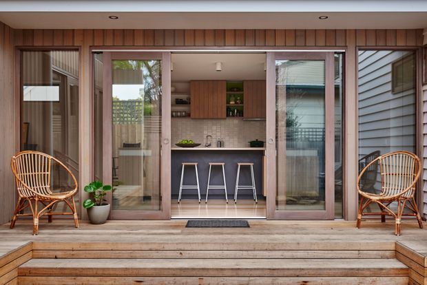 A compact outdoor entertaining area with a small built-in kitchen opening onto a timber deck. The kitchen has warm timber cabinetry, a pale tiled splashback, dark benchtop, open shelving, and three bar stools. Wide timber-framed glazed folding or sliding doors connect the kitchen to the deck, with rattan lounge chairs and potted greenery on either side. Timber-clad walls and broad deck steps create a warm, relaxed setting.