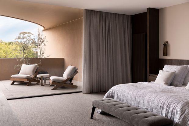 A refined contemporary bedroom with a large upholstered bed, grey tufted bench, dark timber bedside joinery, and full-height grey curtains. The bedroom opens onto a curved glazed sitting area with two lounge chairs and a small side table, overlooking trees. The palette is soft and neutral, with carpeted flooring, warm plaster walls, and a calm, luxurious atmosphere.