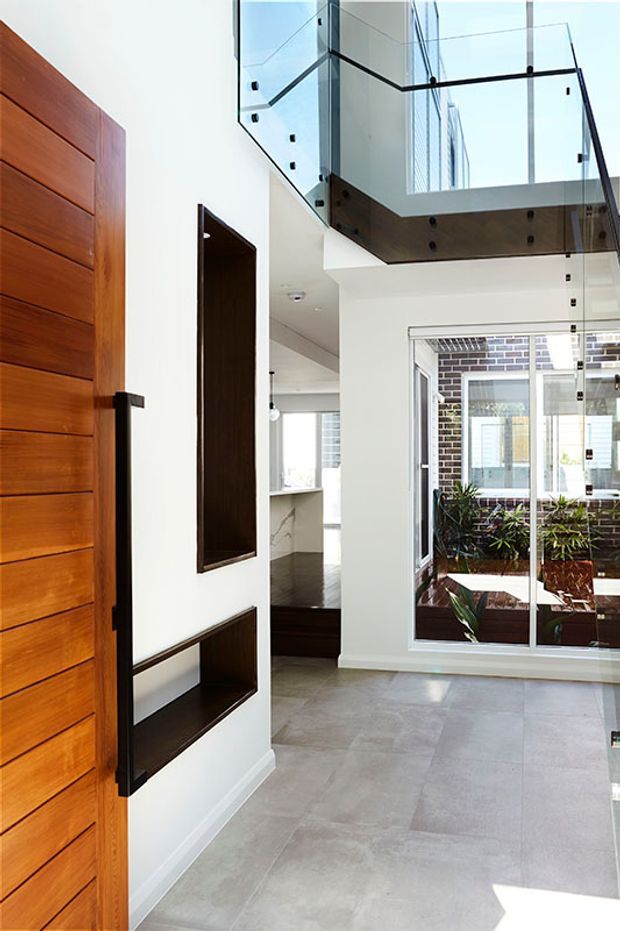 Bright modern entry hall with a large horizontal timber pivot door, white walls, dark built-in display niches, pale stone-look flooring, and a dramatic double-height void with clear glass balustrades. A glazed opening overlooks an internal courtyard or planted lightwell, while a kitchen area is visible beyond.