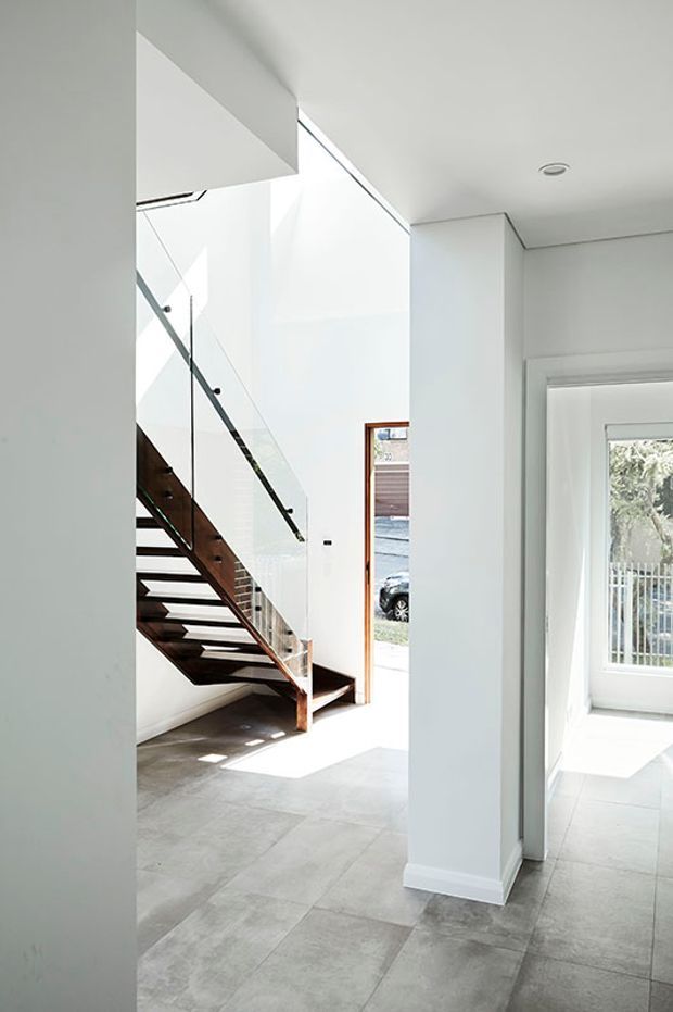 Bright minimalist residential interior showing an open stairway with dark timber treads, a timber stringer, and clear glass balustrade with slim dark handrail. White walls, high ceilings, large glazed openings, and large-format pale grey tiled flooring create an airy entry and circulation space.