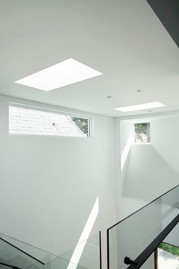 Bright minimalist stairwell or landing with white walls and ceiling, narrow horizontal windows, two rectangular skylights, recessed downlights, and a clear glass balustrade with dark framing. Strong natural sunlight creates geometric light and shadow across the walls.