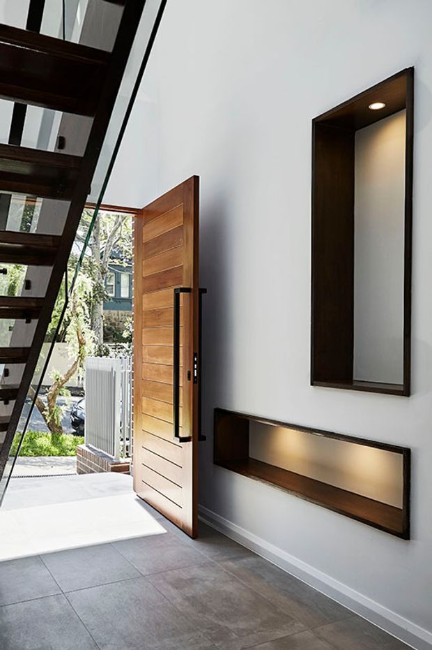 A modern residential entryway viewed from inside, featuring a large timber pivot door opened to the exterior, a floating dark timber stair with glass balustrade, grey tiled flooring, and integrated dark timber wall niches with recessed lighting.